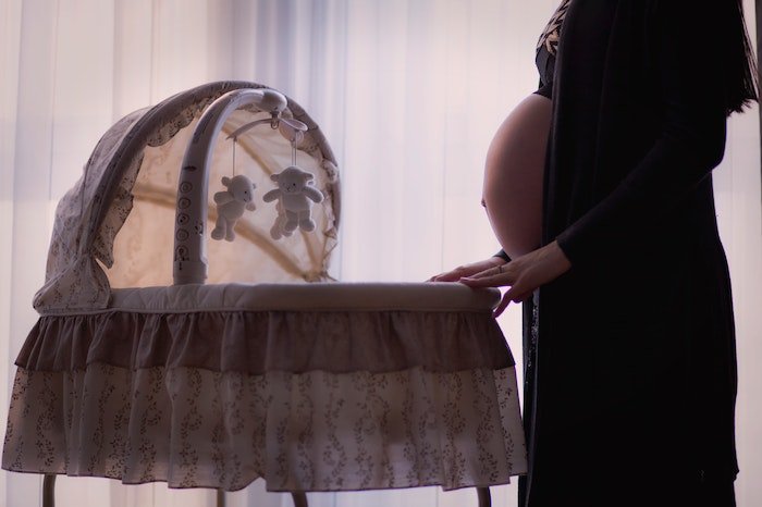 This is an image of a pregnant women standing in front of a bassinet with a canopy. There are 2 bears hanging from the mobiles. Her belly is exposed, and she is wearing a black crop top, and a black cover up.  